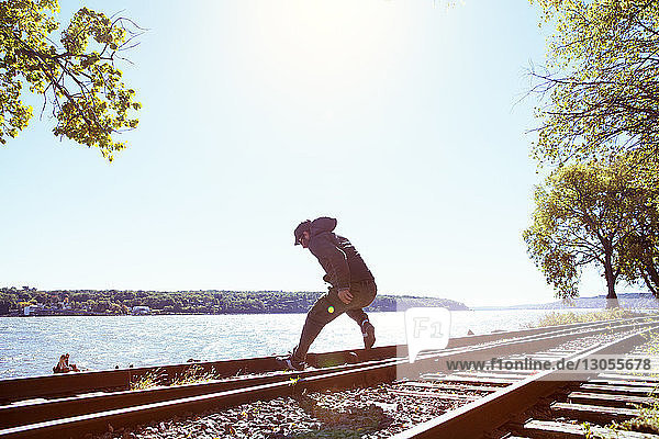 Mann springt auf Eisenbahnschienen am Fluss gegen klaren Himmel