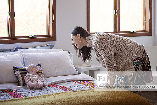 Side view of happy woman looking at cheerful baby girl lying on bed at home