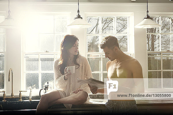Mann liest Zeitung  während seine Freundin mit Kaffeetasse auf dem Küchentisch sitzt