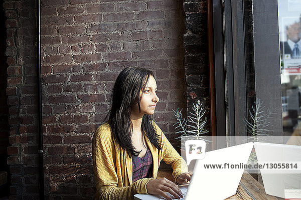 Nachdenkliche Frau benutzt Laptop  während sie zu Hause durchs Fenster schaut