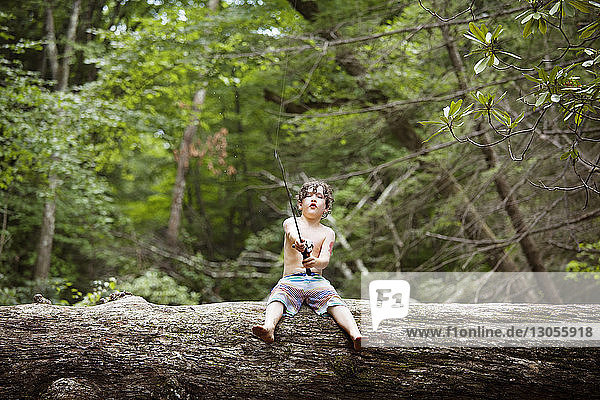 Full length of boy fishing while sitting on fallen tree trunk in forest