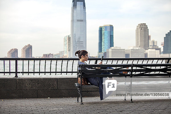 Seitenansicht einer Frau  die einen Tablet-Computer benutzt  während sie auf einer Bank gegen Hudson River sitzt