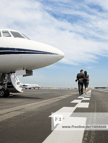 Rear view of business couple walking arms around by corporate jet against sky