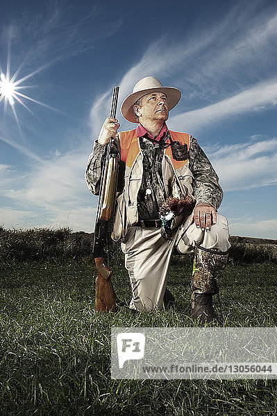 Nachdenklicher älterer männlicher Jäger hält Gewehr auf dem Feld gegen bewölkten Himmel