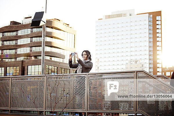 Frau fotografiert mit Smartphone auf Fußgängerbrücke vor Gebäuden