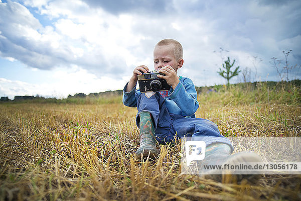 Boy holding camera and sitting on grassy field against sky