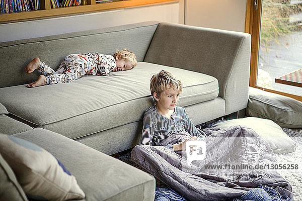 Thoughtful boy sitting by sofa while brother playing in home