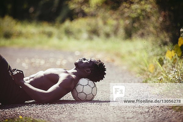 Seitenansicht eines Mannes ohne Hemd  der sich an einem sonnigen Tag auf einem Fussball ausruht