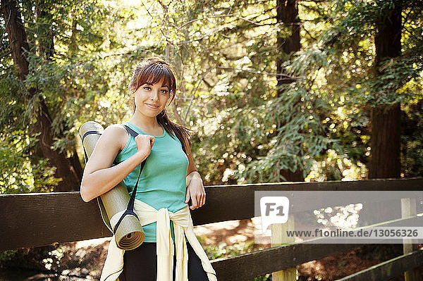 Porträt einer selbstbewussten Frau  die eine Übungsmatte trägt  während sie im Wald an einem Geländer steht