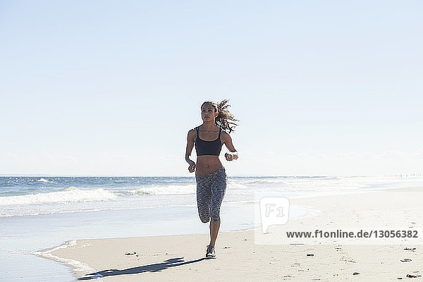 Zuversichtliche Frau joggt am Strand am Strand gegen den Himmel