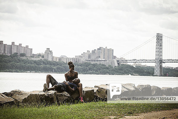 Couple relaxing on rocks at riverbank against bridge