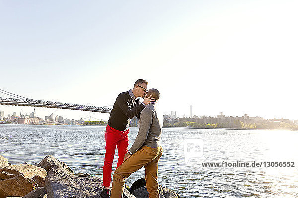 Man kissing boyfriend while standing on rocks by East River on sunny day