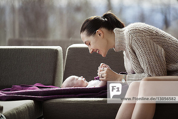 Side view of happy woman playing with baby girl on sofa at home