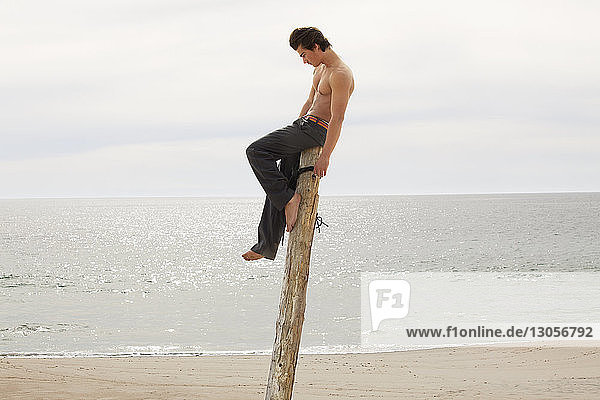 Shirtless man sitting on wooden post at beach