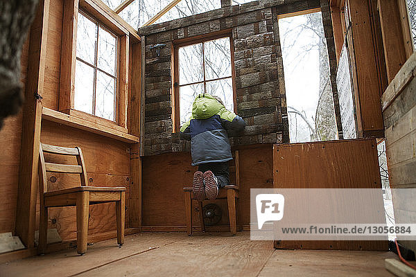 Rückansicht eines Jungen  der durch ein Fenster schaut  während er auf einem Stuhl im Baumhaus kniet