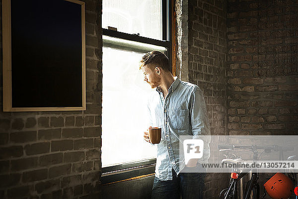 Mann mit der Hand in der Tasche hält Kaffeetasse  während er zu Hause am Fenster steht
