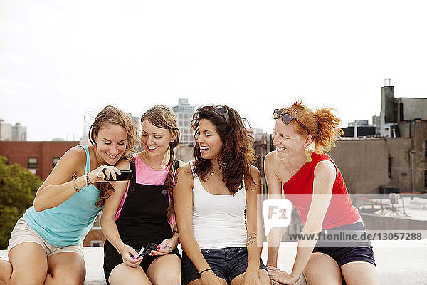Lächelnde Frauen schauen auf ihr Handy  während sie auf der Gebäudeterrasse vor klarem Himmel sitzen