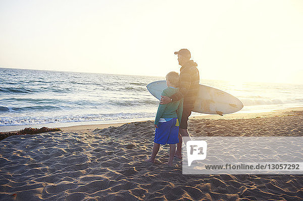 Älterer Mann hält Surfbrett  während er mit seinem Enkel am Strand steht