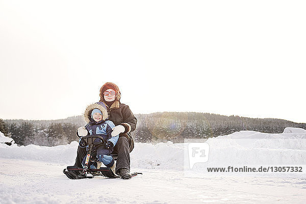 Portrait of happy mother and son sitting on sled during winter against clear sky