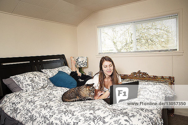 Glückliche Frau  die einen Laptop benutzt  während sie neben einer Katze auf dem Bett liegt