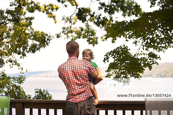 Rückansicht eines Vaters  der einen Sohn trägt  während er am Geländer am See steht