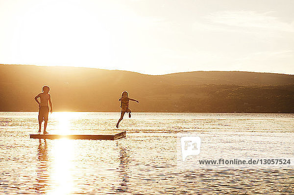 Schwester taucht in den See  während der Junge bei Sonnenuntergang auf einer schwimmenden Plattform steht