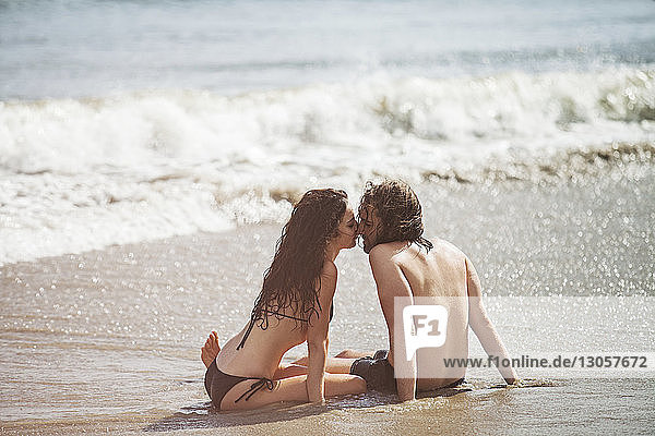 Romantisches Paar reibt sich während der Sommerferien am Strand die Nase  während es am Ufer sitzt
