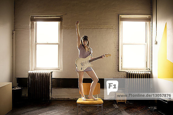 Glückliche Frau spielt Gitarre  während sie zu Hause auf einem Stuhl steht