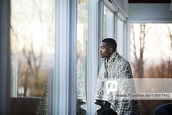 Mann in Decke gehüllt  schaut weg  während er zu Hause am Fenster steht