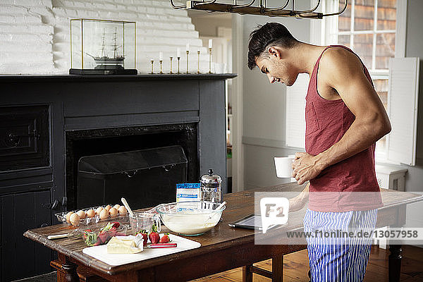 Mann mit Kaffeetasse  der einen Tablet-Computer benutzt  während er in der Küche am Tisch steht