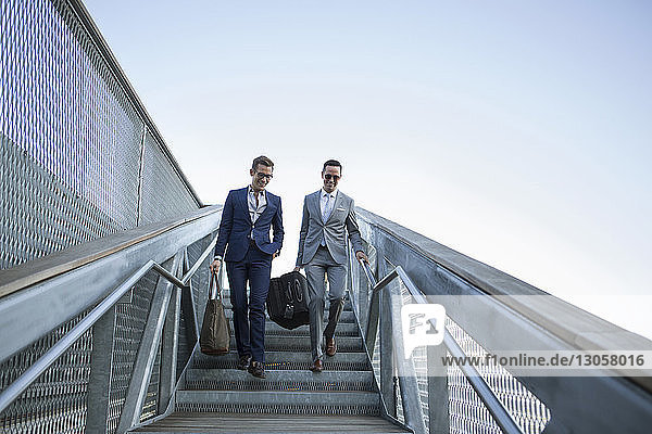 Geschäftsleute tragen Gepäck  während sie sich auf einer Treppe gegen den klaren Himmel nach unten bewegen