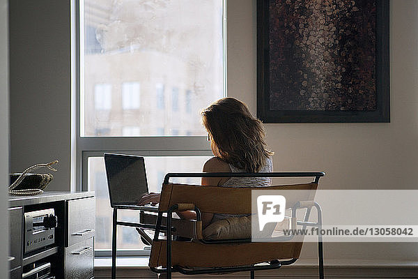 Rückansicht einer Frau  die einen Laptop benutzt  während sie zu Hause auf einem Stuhl sitzt