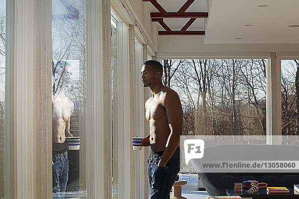 Mann ohne Hemd hält Becher  während er zu Hause durchs Fenster schaut