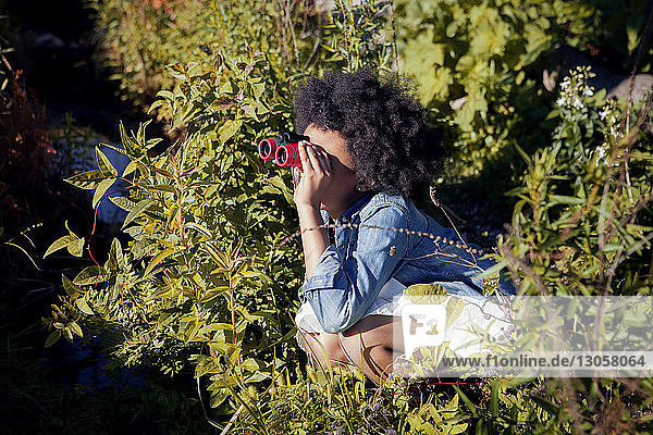 Mädchen kauert inmitten von Pflanzen und schaut durch ein Fernglas