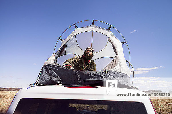 Low angle view of thoughtful man sitting in tent on car roof against blue sky