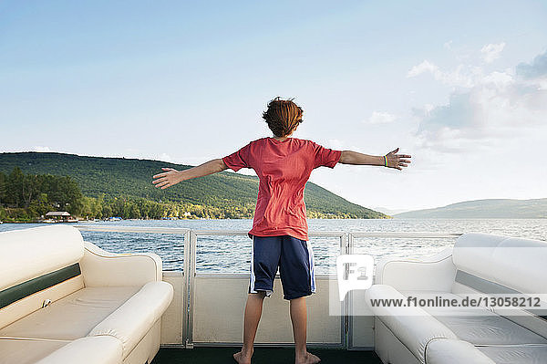 Rear view of boy with arms outstretched standing in boat on lake against sky