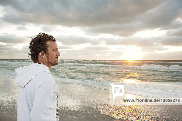 Side view of thoughtful man standing on beach against cloudy sky at sunset