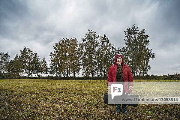 Portrait of female farm worker holding buckets while standing on field against cloudy sky