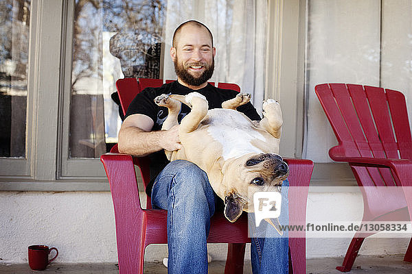 Mann spielt mit Hund  während er auf einem Stuhl auf der Veranda sitzt