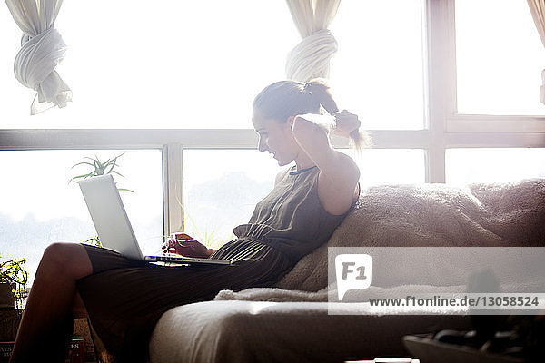 Frau benutzt Laptop-Computer  während sie zu Hause auf dem Sofa am Fenster sitzt