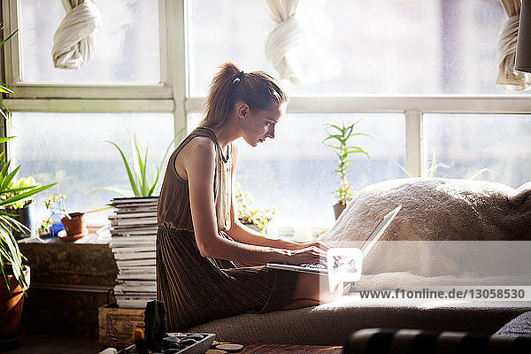 Frau benutzt Laptop-Computer  während sie zu Hause auf dem Sofa am Fenster kniend sitzt