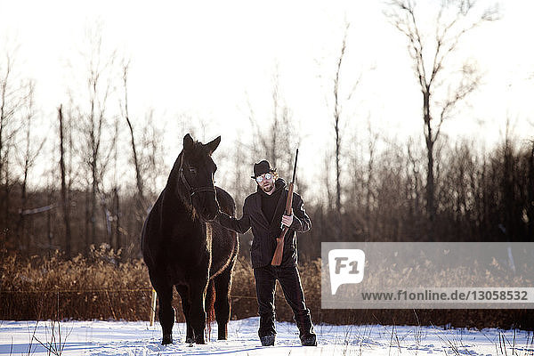 Mann hält Gewehr stehend mit Pferd auf schneebedecktem Feld