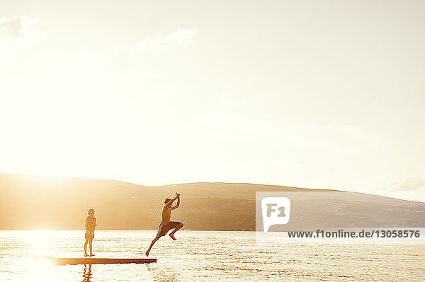 Bruder taucht in den See  während ein Mädchen bei Sonnenuntergang auf einer schwimmenden Plattform gegen den Himmel steht