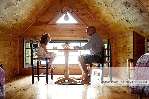 Seitenansicht eines Vaters  der mit seiner Tochter am Tisch in einem Holzhaus sitzt