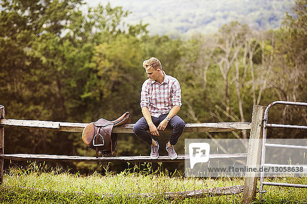 Mann schaut auf Sattel  während er auf einem Geländer in einem Bauernhof sitzt
