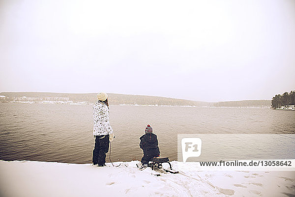 Mutter steht bei ihrem Sohn auf einem Schlitten am Seeufer vor klarem Himmel