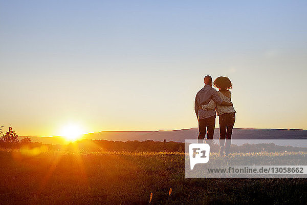 Rückansicht eines Paares,  das bei Sonnenuntergang auf Gras vor klarem Himmel steht
