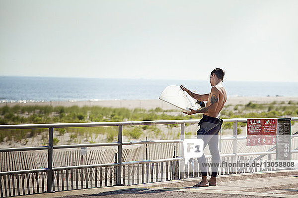 Mann sieht Surfbrett an  während er auf der Brücke gegen das Meer steht