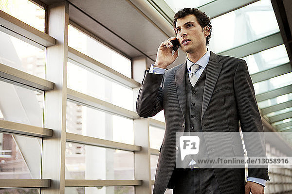 Niedrigwinkelansicht eines Geschäftsmannes  der mit einem Mobiltelefon spricht  während er auf einem überdachten Gehweg geht