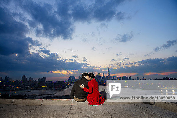 Rückansicht eines jungen Paares  das sich in der Abenddämmerung gegen Fluss und Stadtbild küsst
